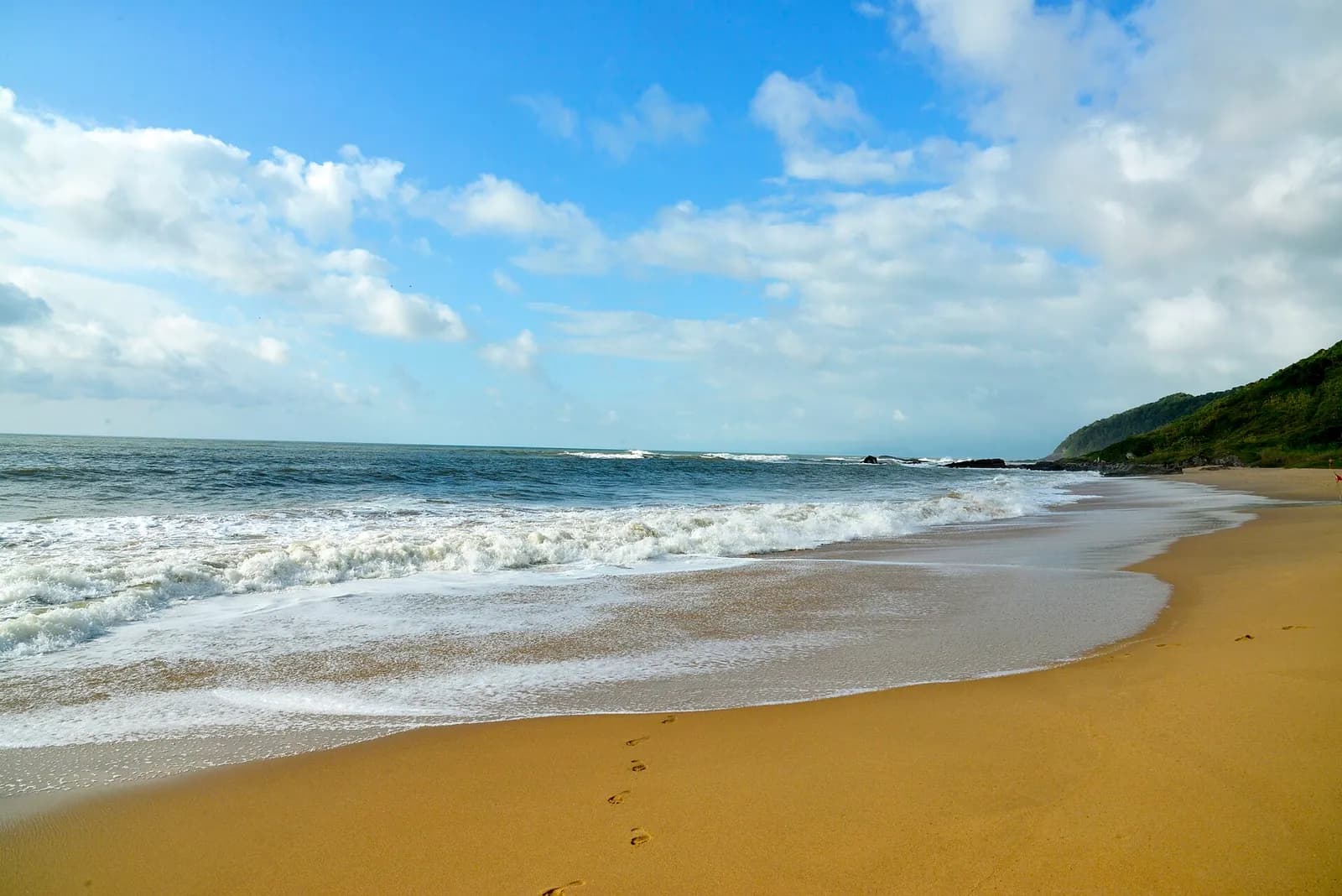 Ondas quebrando na areia dourada da Praia Vermelha em Penha, com morro verde ao fundo