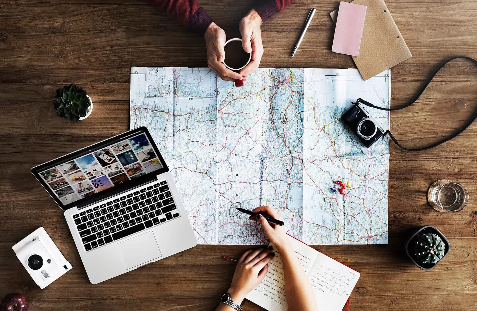 Mãos sobre uma mesa de madeira, planejando viagem com mapa, laptop e café